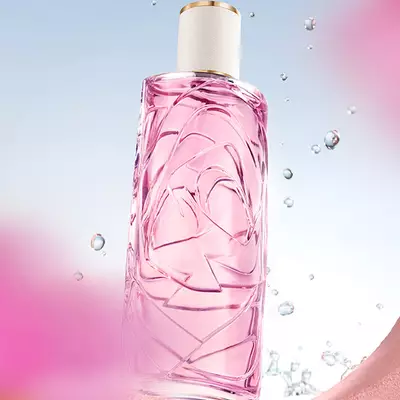 A light pink perfume bottle with a textured glass design, against a light blue sky background, with water droplets splashing around it. The bottle is seen through a circular opening in a textured pink wall, and there are blurred pink flowers in the foreground and background.