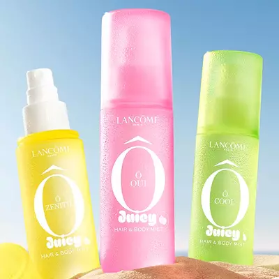 Two green bottles, one perfume rectangular bottle with a textured design and one spray bottle, standing on a sandy beach. A folded pink striped beach towel lies next to the bottles. In the background, there is a clear blue sky and the ocean.