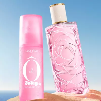 A beach scene with three colorful perfume bottles standing upright in the sand, one pink, one yellow, and one green, with another pink perfume bottle lying on its side in the foreground. A pink and white striped towel is draped over the sand to the left. The background features a clear blue sky transitioning to a blue ocean at the horizon.